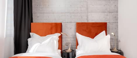 Cama doble con ropa de cama naranja y almohadas blancas, mesita de noche negra con lámpara, y pared de concreto al fondo.