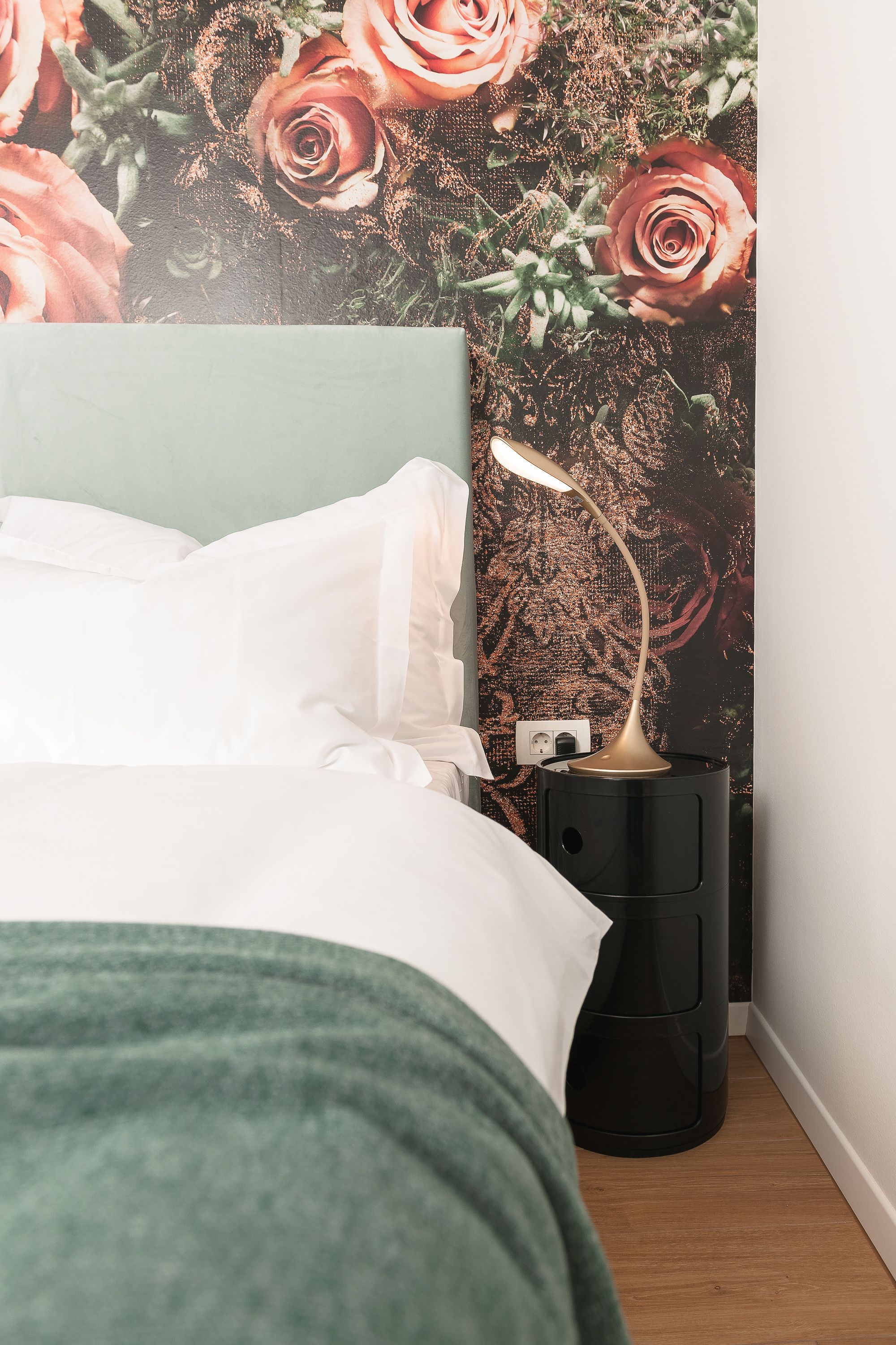 Un lit bien fait, avec du linge de lit blanc, un plaid vert et un mur recouvert d’un papier peint à motifs floraux. Une table de chevet noire avec une lampe se trouve à côté du lit.