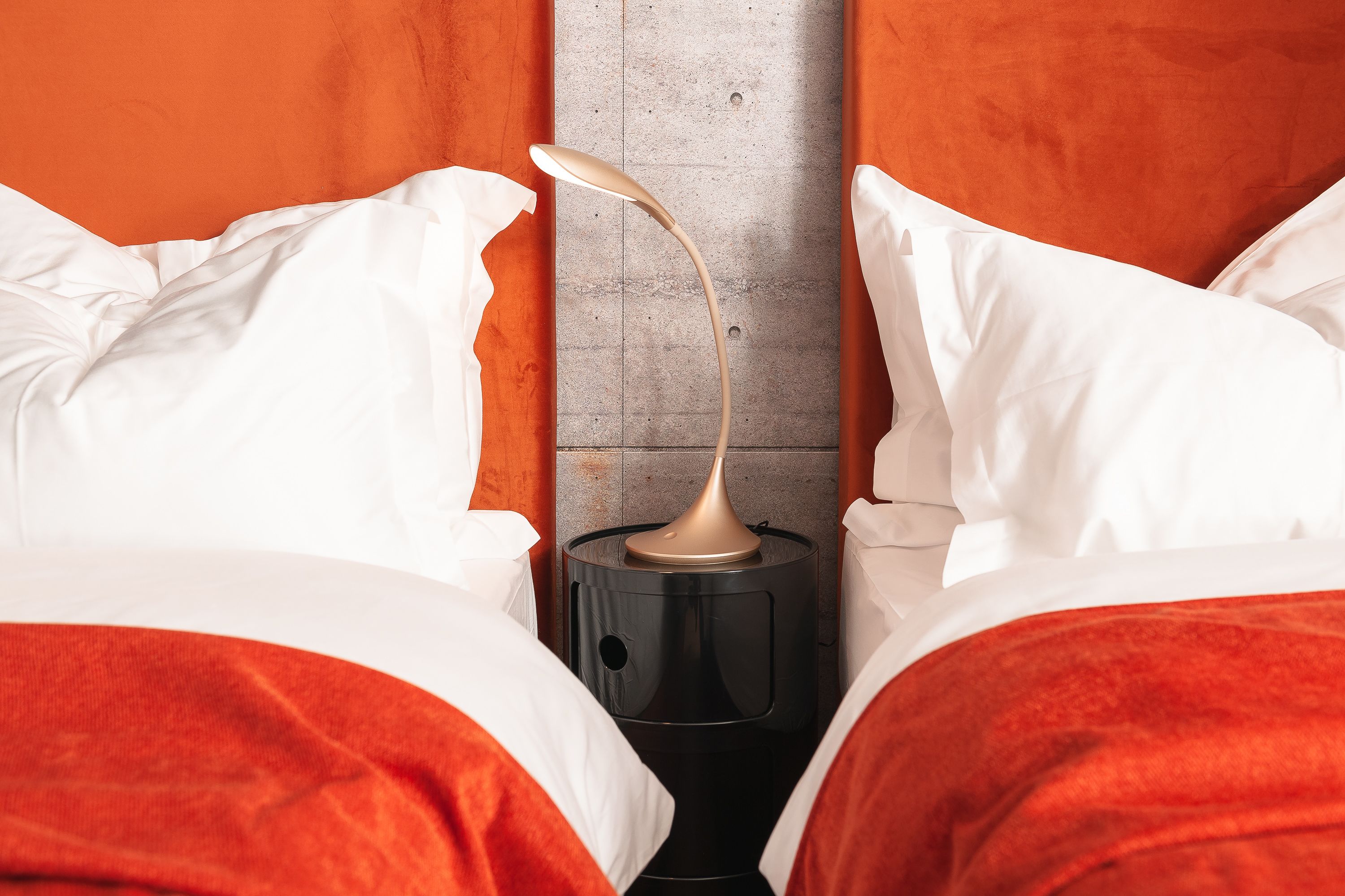 A bed with white and red bedding, a black nightstand with a lamp, and a grey wall.