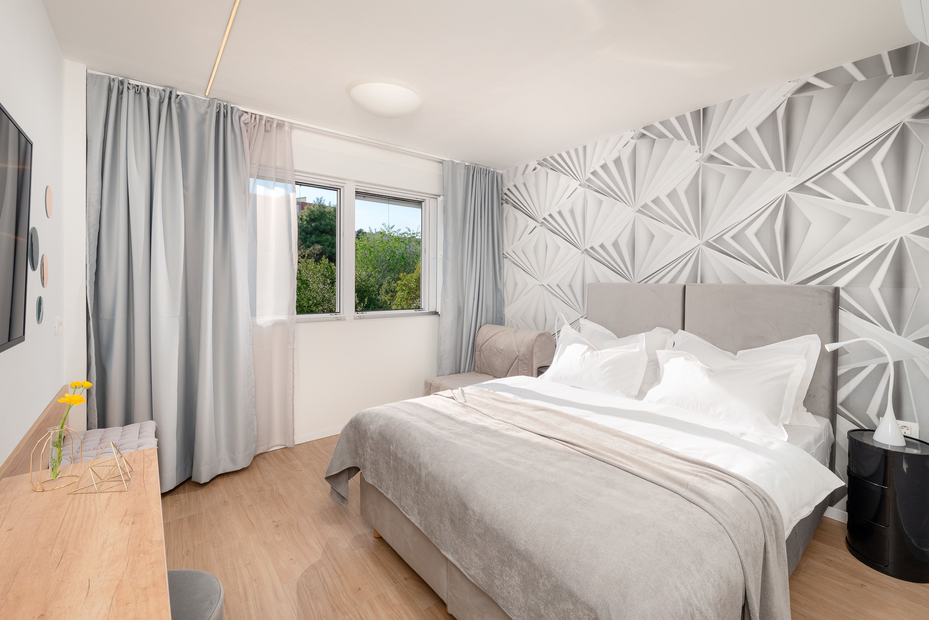 Une chambre moderne avec un grand lit, une table de chevet, une fenêtre avec des rideaux et un motif géométrique sur le mur.