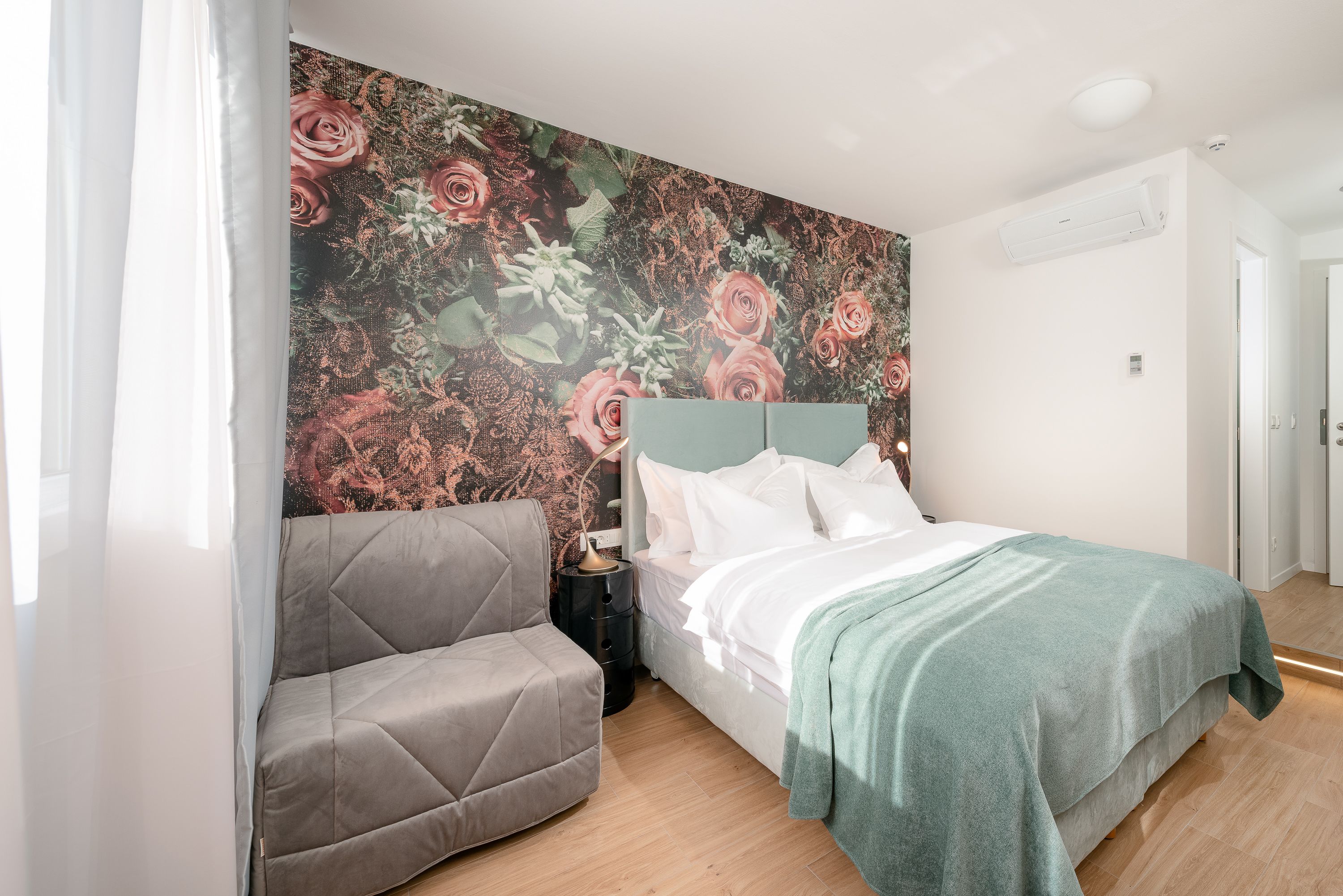 Une chambre à coucher avec un lit, un fauteuil gris, une table de chevet et un papier peint à motifs floraux.