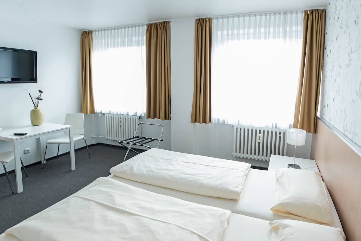 Quarto de hotel com duas camas, uma escrivaninha, uma cadeira, um radiador e uma TV fixada na parede.