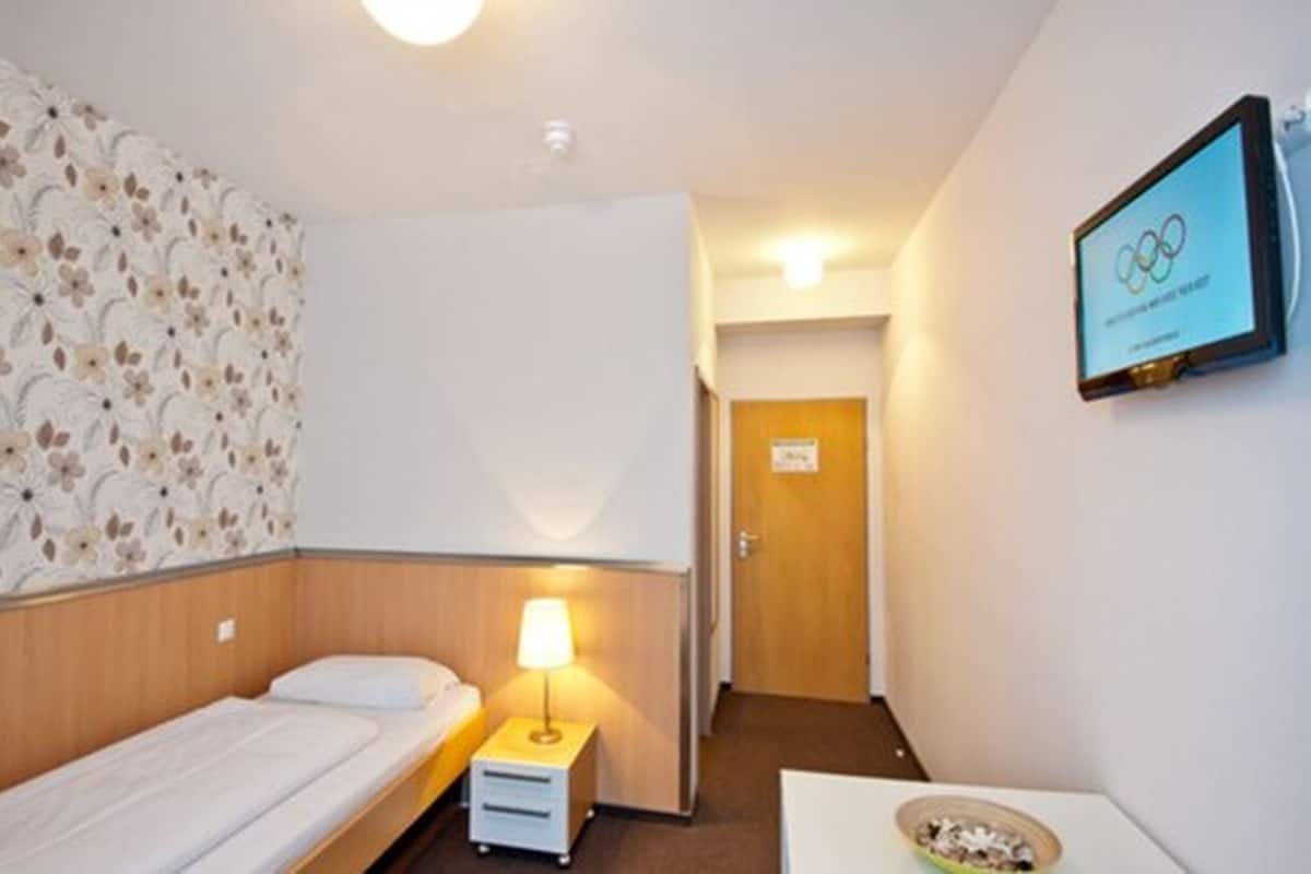 Quarto de hotel com uma cama de solteiro, uma mesa de cabeceira com luminária, televisão na parede e papel de parede estampado com flores.