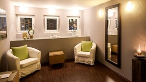 Ein Zimmer mit zwei weißen Sesseln, einem kleinen Holztisch, einem Spiegel und einem Fenster mit Jalousien.