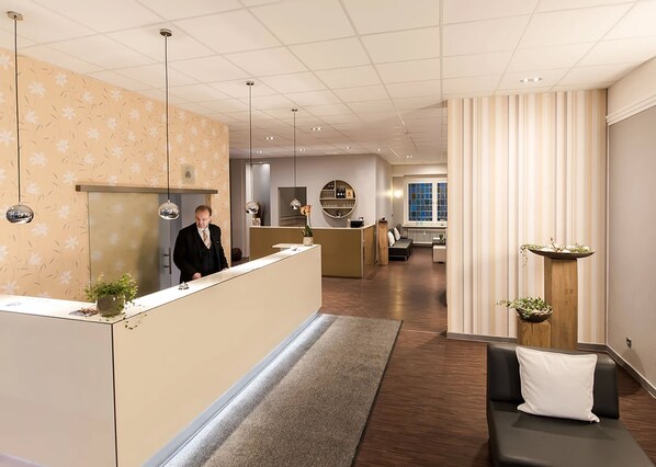 A hotel lobby with a reception desk, a seating area with a sofa and a small table, and a kitchen area in the background.