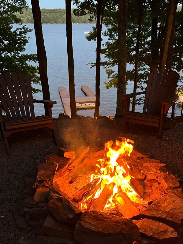 A campfire with two Adirondack chairs facing a lake.