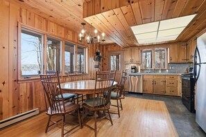A wooden cabin interior with a dining table, chairs, a chandelier, and a kitchen area.