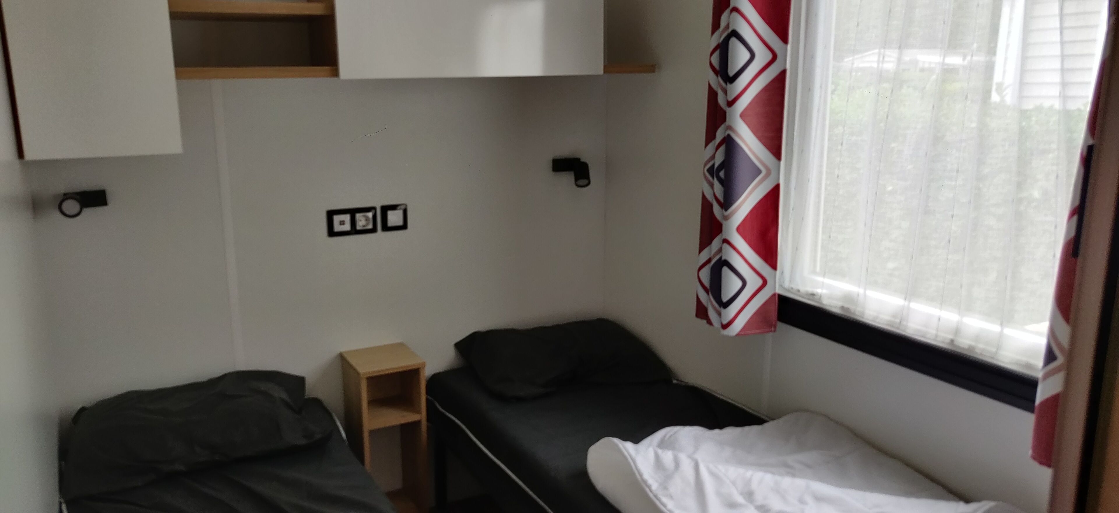 A small, compact room with two single beds, a small wooden shelf, and a window with a patterned curtain.