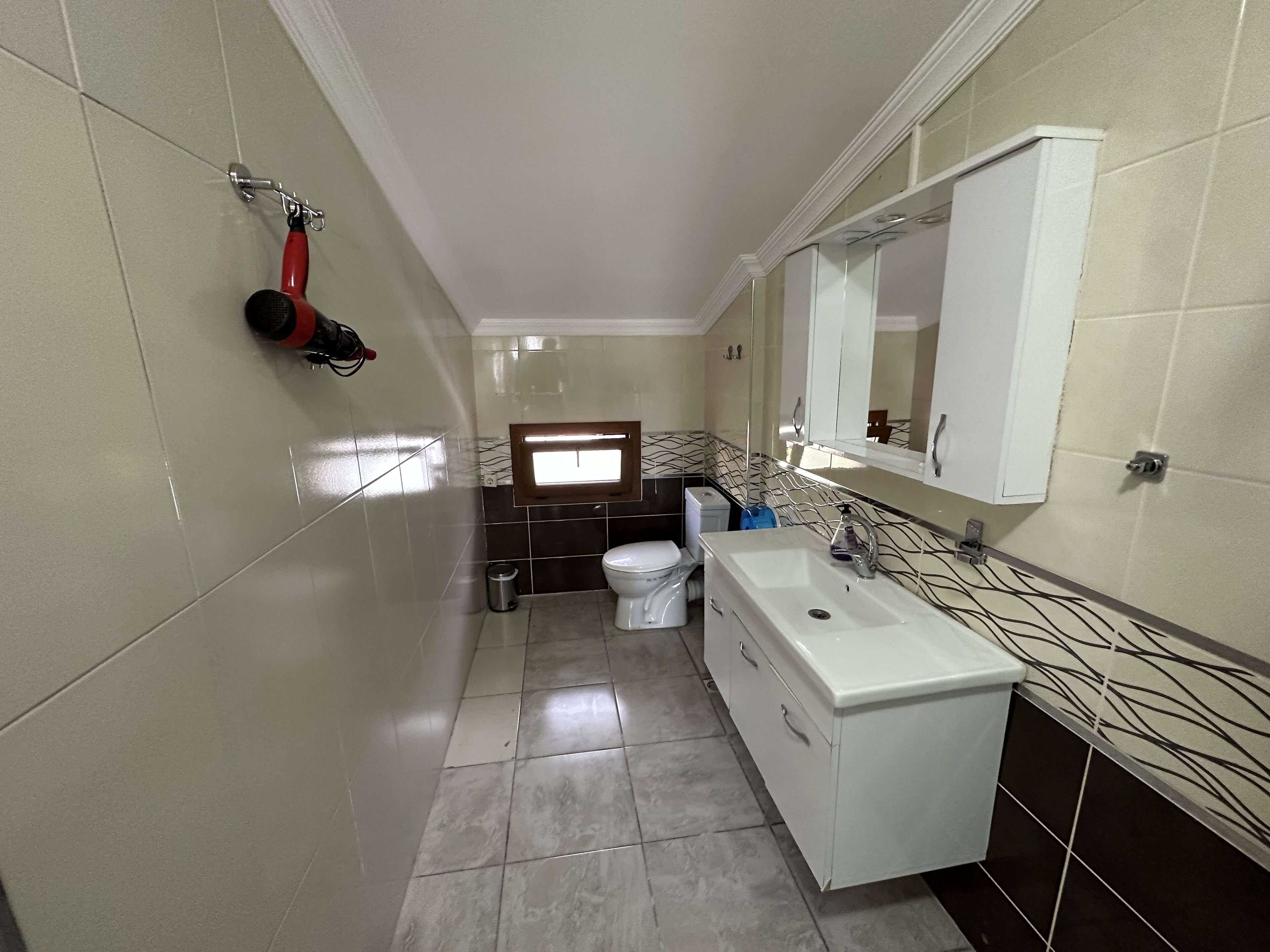 Une salle de bain avec un lavabo blanc, un miroir et des toilettes. Une fenêtre laisse entrer la lumière naturelle.