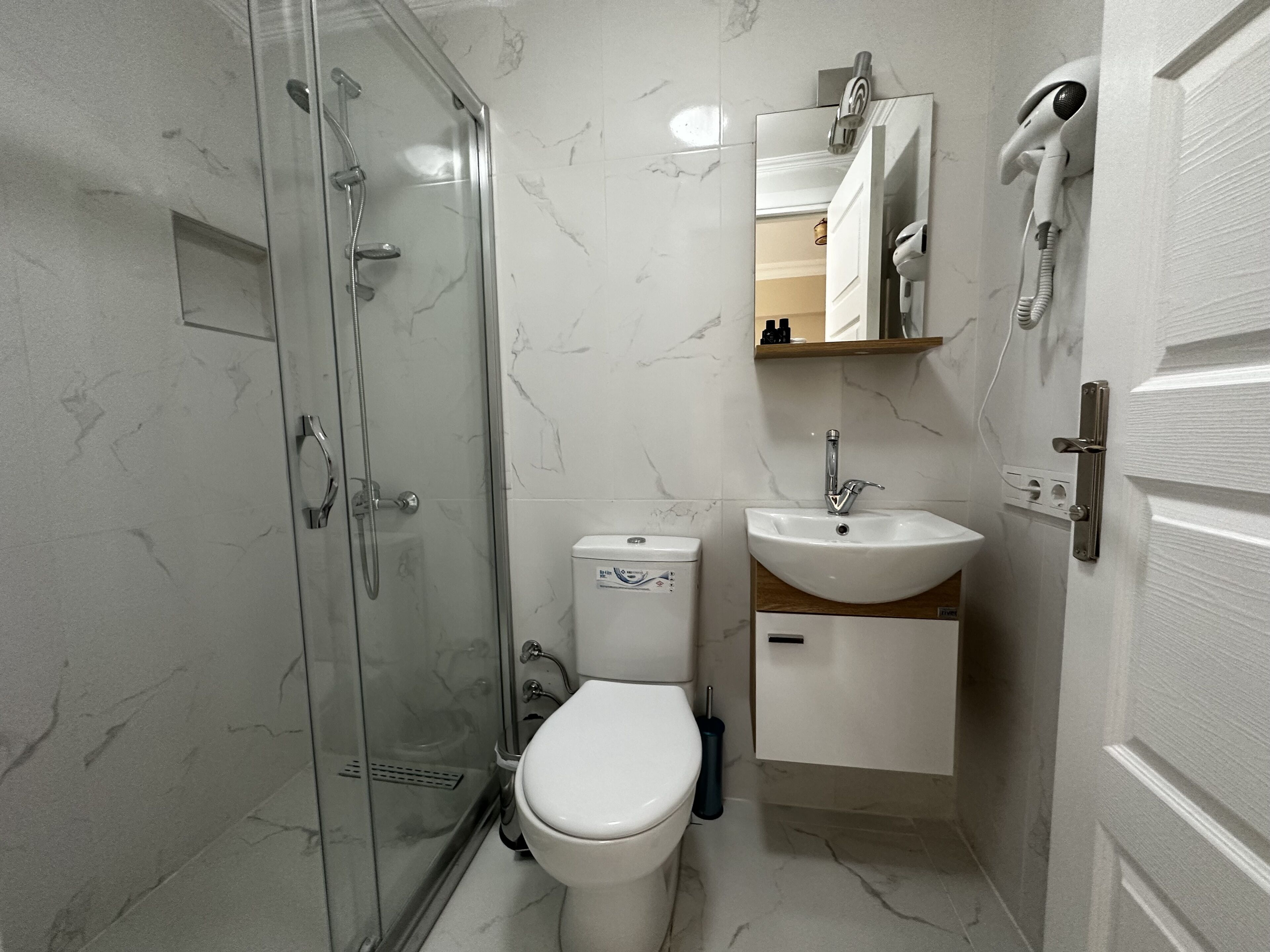 A bathroom with a glass-enclosed shower, a toilet, a sink, and a hairdryer mounted on the wall.