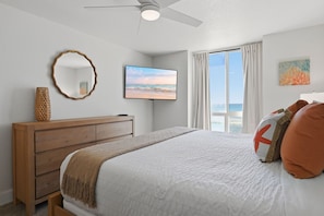 A bedroom with a bed, a wooden dresser, a television, a ceiling fan, and a window with curtains.