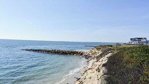 A coastal scene with a sandy beach, a wooden walkway, and a house in the background. - Seven bedroom directly across street from shared/private beach (Dennis Port)