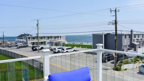 A view from a balcony overlooking a street with parked cars, a building with a balcony, and a body of water in the background. - Seven bedroom directly across street from shared/private beach (Dennis Port)