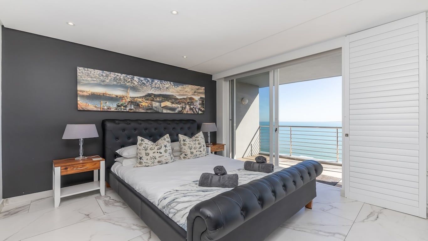 A modern bedroom with a large bed, a painting, a nightstand, and a view of the sea.