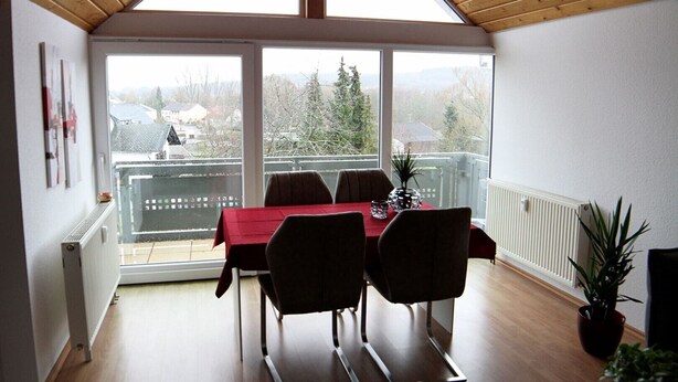 Un coin repas avec une table rouge et quatre chaises, une plante en pot, et une vue sur les arbres et les bâtiments à travers de grandes fenêtres.