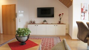 Un salon avec un téléviseur à écran plat fixé au mur, un meuble blanc orné d’objets décoratifs, un tapis rouge et une plante dans un pot noir.