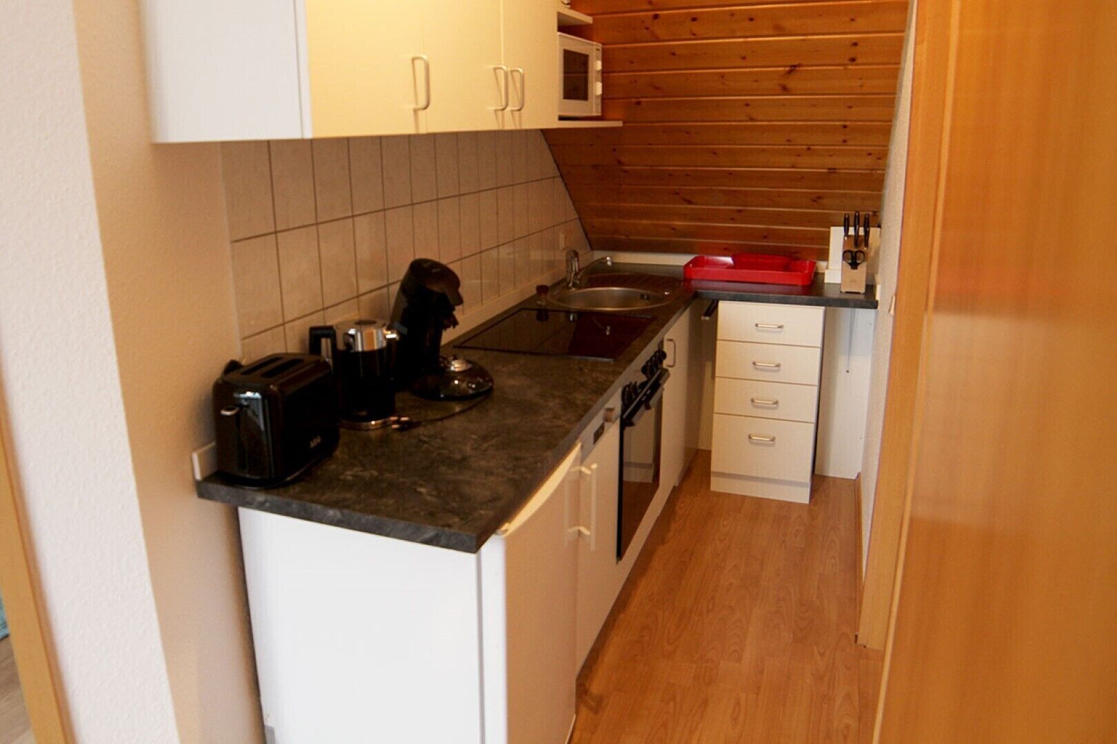A compact kitchen with white cabinets, a microwave, a coffee maker, and a red dish rack.