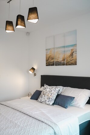 Une chambre à coucher bien rangée, avec un lit, des oreillers, une lampe fixée au mur et un tableau représentant un paysage.