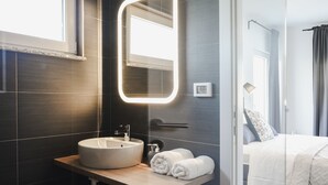 A modern bathroom with a round sink, a mirror with integrated lighting, and a view into a bedroom with a bed and curtains.