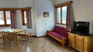 Una habitación con una mesa y sillas de madera, un sofá, un televisor sobre un mueble de madera y ventanas grandes con cortinas.