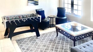A room with a wooden ceiling, a ceiling fan, a foosball table, a glass-top coffee table, a sofa, and a window with a view. - Chateau Saguaro is your home away from home (La Quinta)