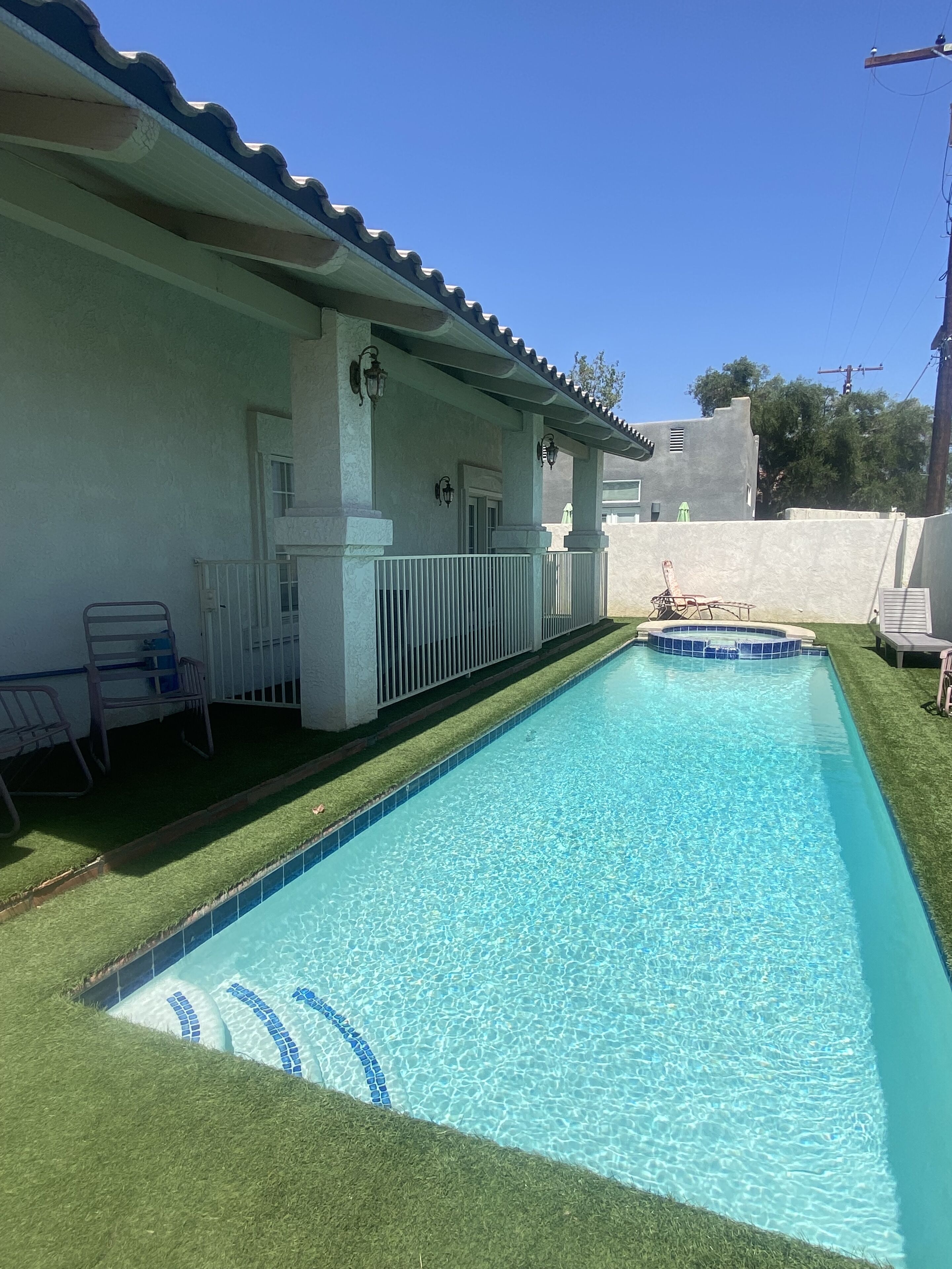 A rectangular swimming pool with a tiled edge and a small hot tub area, surrounded by a white building with a tiled roof and a fenced patio area with chairs.