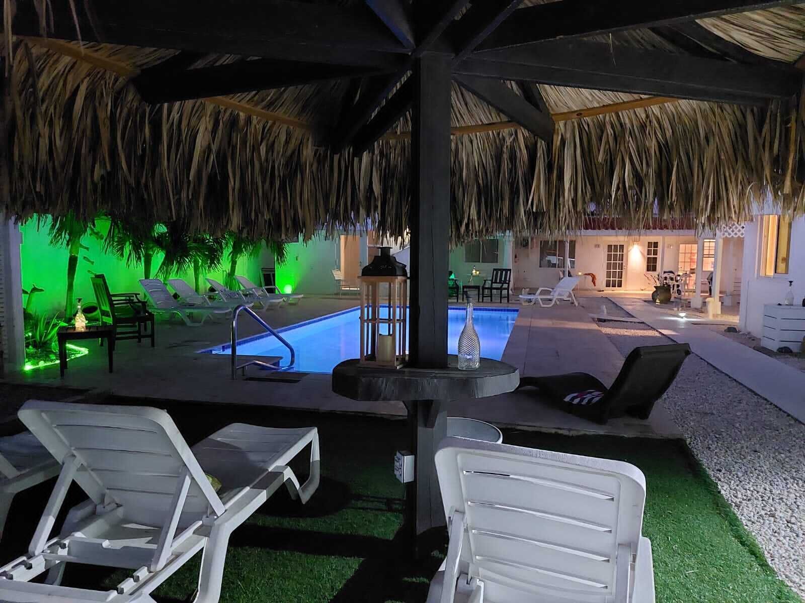 Un espace piscine avec des chaises longues, un toit de chaume et une petite table sur laquelle se trouve une bouteille.