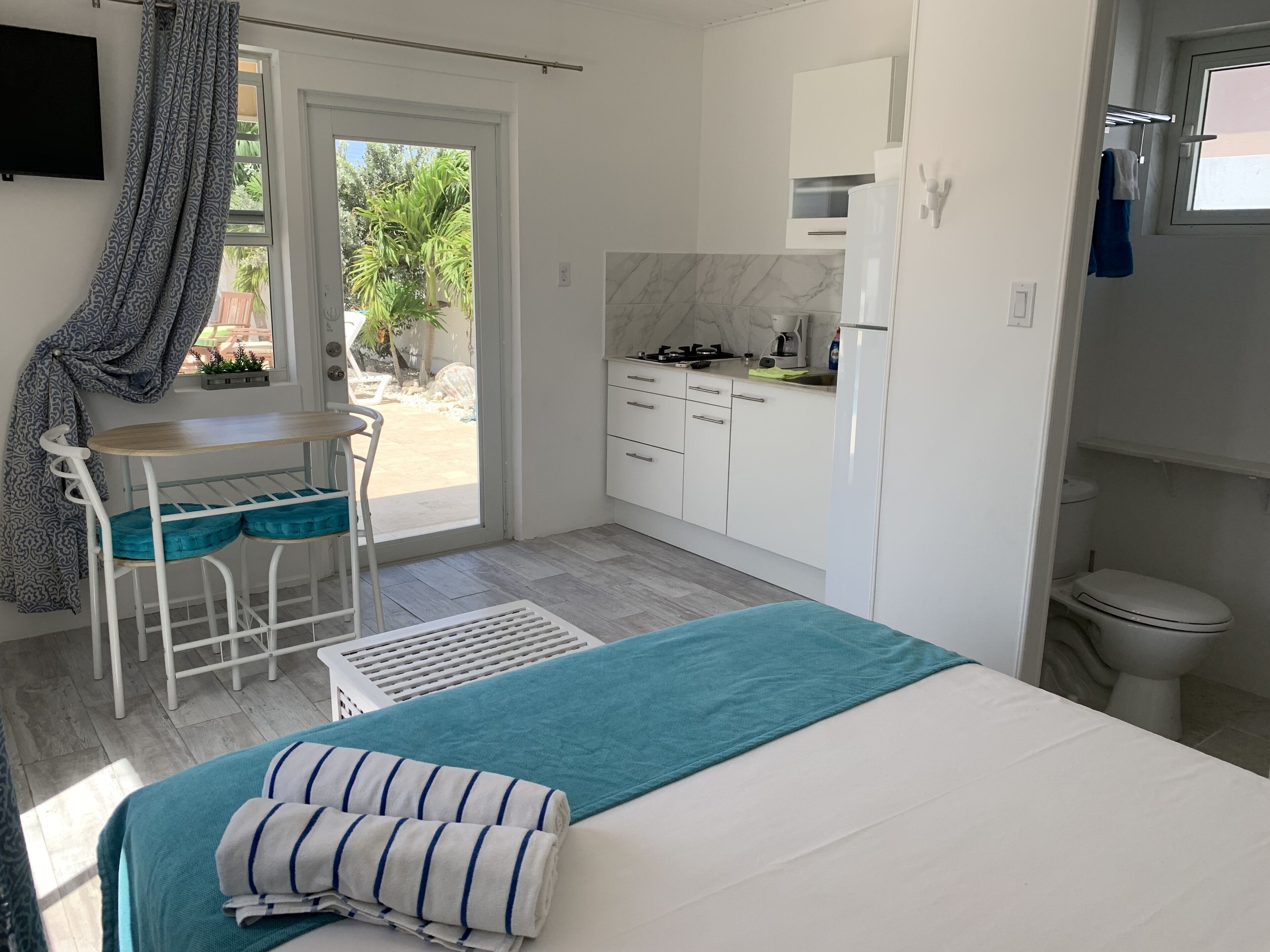 A compact bedroom with a bed, a small table, and a chair. A kitchen area is visible with a sink, stove, and cabinets.