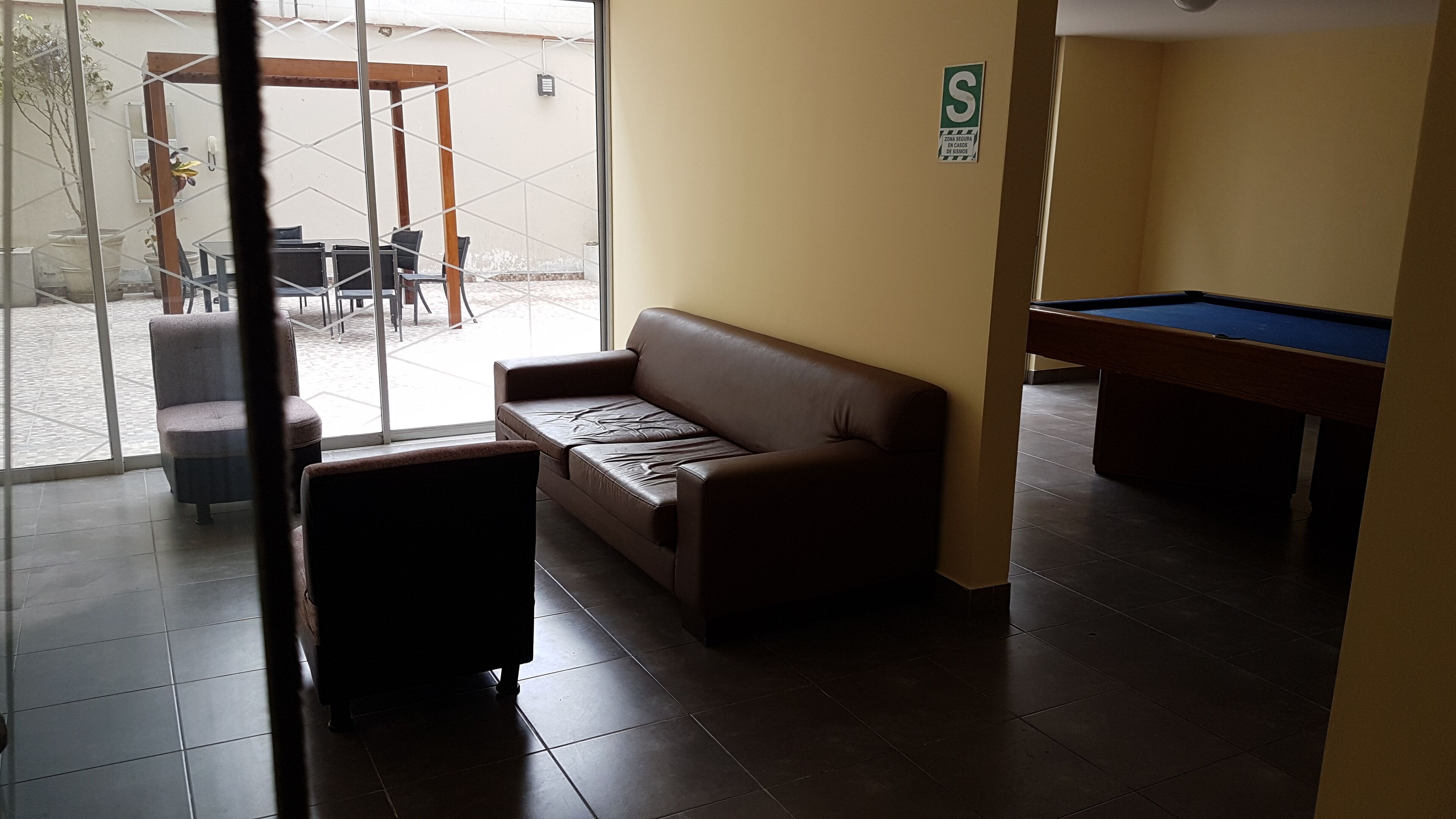 A room with a brown sofa, a pool table, and a glass door leading to an outdoor area with tables and chairs.