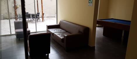 A room with a brown sofa, a pool table, and a glass door leading to an outdoor area with tables and chairs.