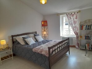 Une chambre à coucher comprenant un lit, une table de chevet, une lampe, une fenêtre avec des rideaux, un radiateur et une bibliothèque.