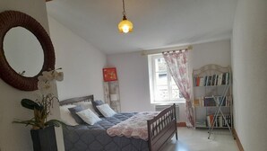 Un dormitorio con una cama, una mesita de noche, un espejo, una ventana con cortinas, una estantería y una lámpara colgante.