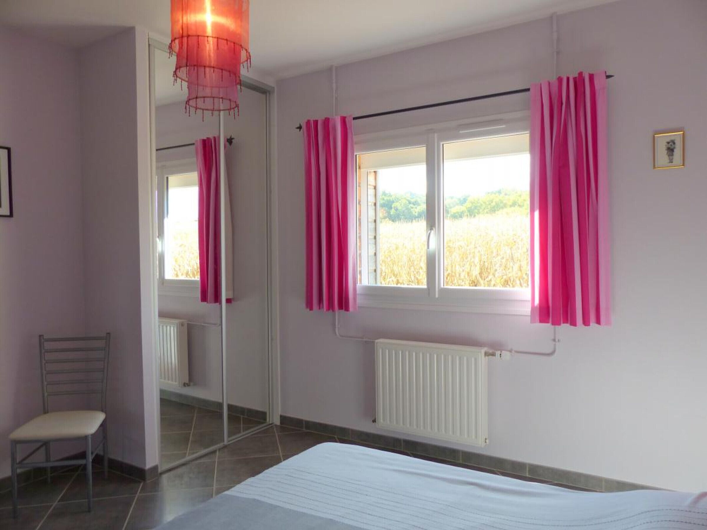 Une chambre à coucher avec un lit, une chaise, une fenêtre avec des rideaux roses et un radiateur.