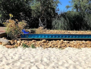 Relax by the serene pool surrounded by lush greenery and a sandy beach area.
