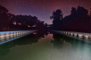 Een nachtelijke scène met een sterrenhemel, weerspiegeld in kalm water, omringd door bomen en een brug.