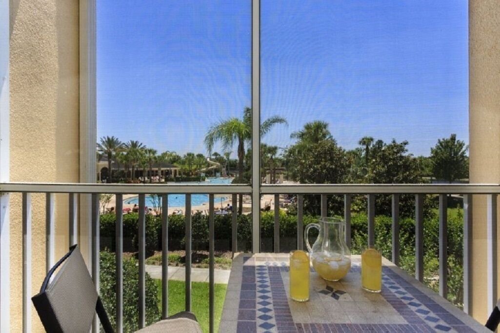 A balcony with a table, two glasses of a yellow beverage, and a view of a pool and palm trees.
