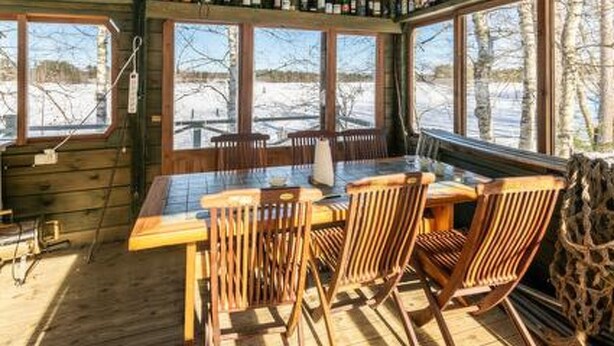 A wooden cabin with a dining area featuring a table and chairs, large windows with a view of a snowy landscape, and shelves with bottles.