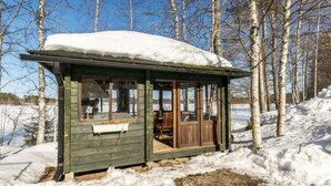 A small wooden cabin with a snow-covered roof, a porch with a bench, and a view of a snowy landscape with trees.