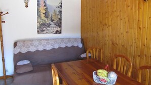 A wooden dining table with a basket of fruit, a bench with a lace cover, and a painting of a forest scene on the wall. - Charming T3 "Les Cerdans" 4 seasons 2 people (Porté Puymorens)