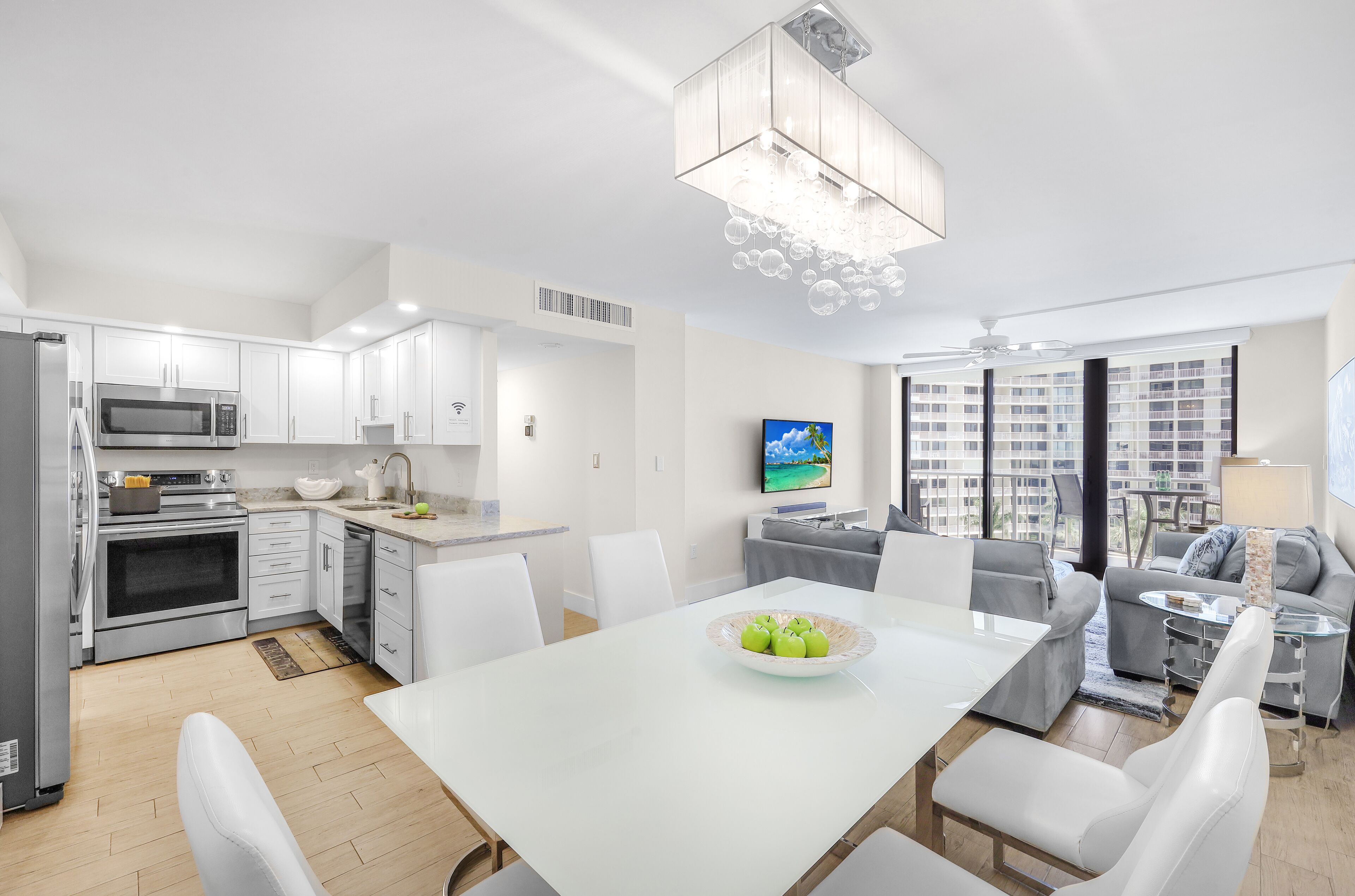 A modern kitchen with white cabinetry, a dining area with a white table, and a living space with a grey sofa and a TV mounted on the wall.