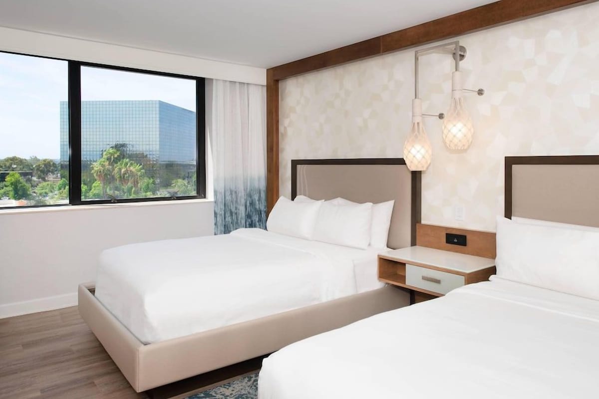 Habitación de hotel con dos camas, un ventanal grande con vistas a la ciudad y una lámpara moderna.