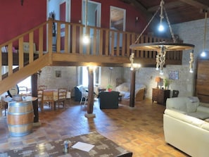 Un loft spacieux avec un escalier en bois, un luminaire suspendu et différents coins salon, notamment un canapé et des fauteuils.