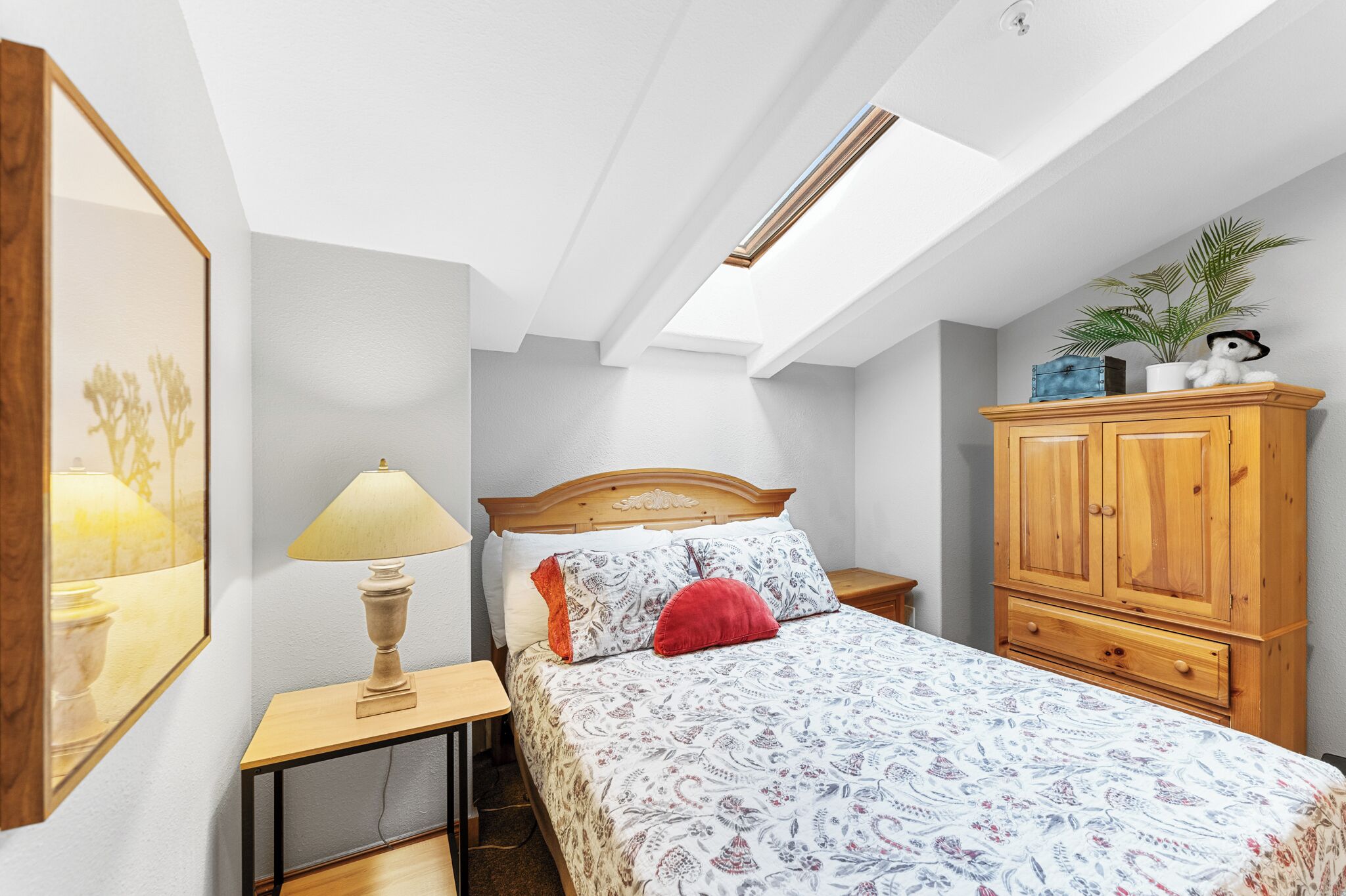 A bedroom with a skylight, a bed with patterned bedding, a wooden nightstand, a lamp, a red pillow, a wooden wardrobe, and a framed picture on the wall.