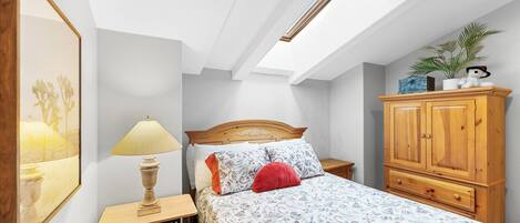 A bedroom with a skylight, a bed with patterned bedding, a wooden nightstand, a lamp, a red pillow, a wooden wardrobe, and a framed picture on the wall.