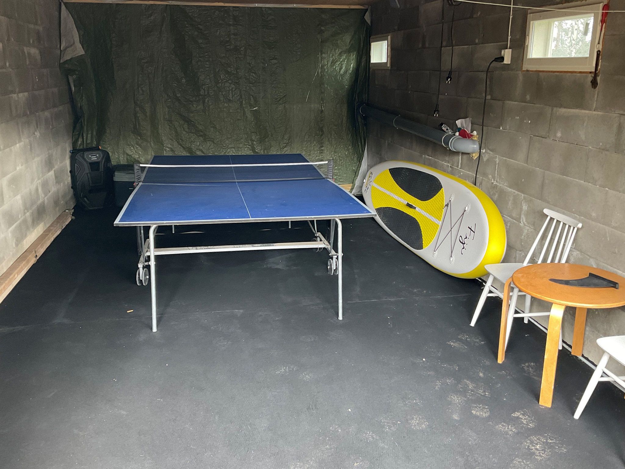Une table de ping-pong dans une pièce dont le sol est en béton, avec une raquette jaune et blanche, et une petite table en bois accompagnée d’une chaise.