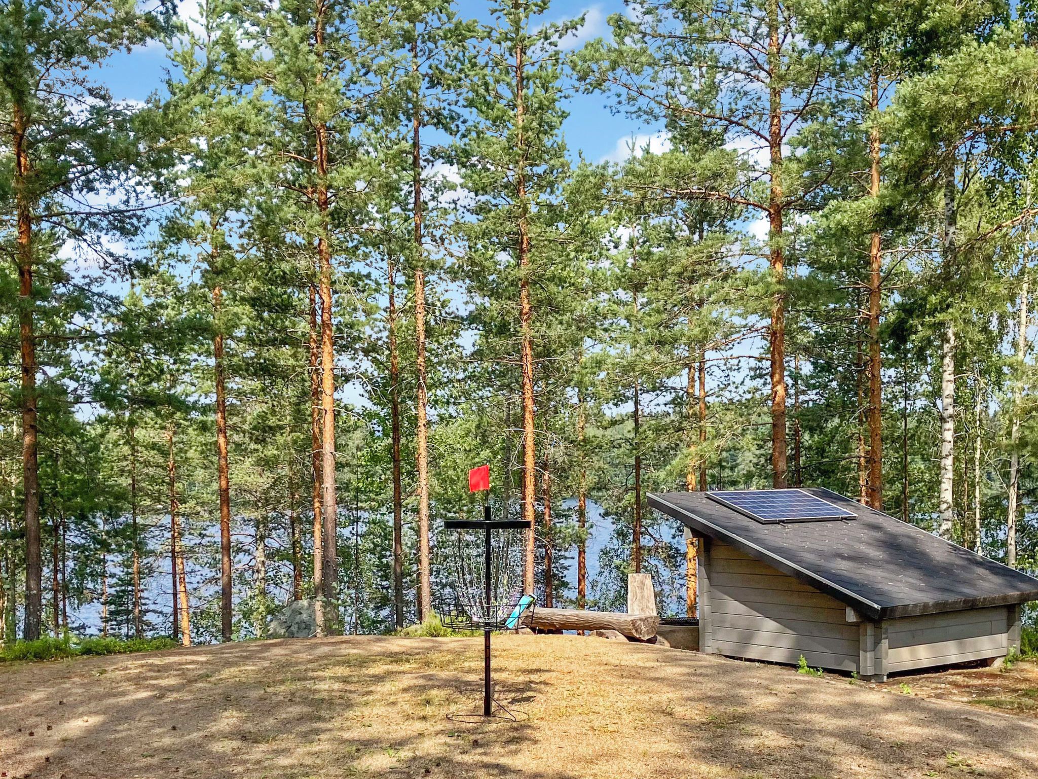 Une cabane surmontée d’un toit équipé de panneaux solaires, une structure métallique avec un drapeau rouge, et une zone boisée avec un lac en arrière-plan.