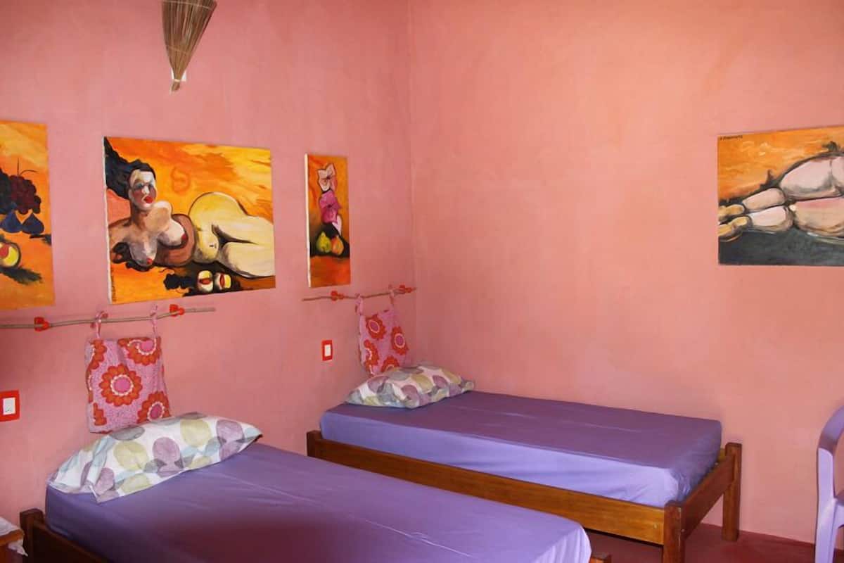 Une chambre avec deux lits, des murs roses et trois tableaux.