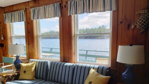 A wooden cabin interior with a striped sofa, two lamps, and large windows overlooking a body of water. - Waterfront Lake House Cottage Private Dock, Deck, Kayak, Boat, Swim, Fish, RELAX (Deerfield)