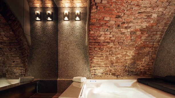 A spa room with a brick wall, a wooden floor, a large bathtub, and two wall-mounted lights.
