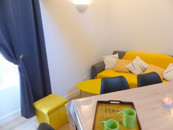 Et værelse med en gul sofa, en lille gul skammel, et træbord med en bakke og to grønne krus, samt en mørkeblå gardin.